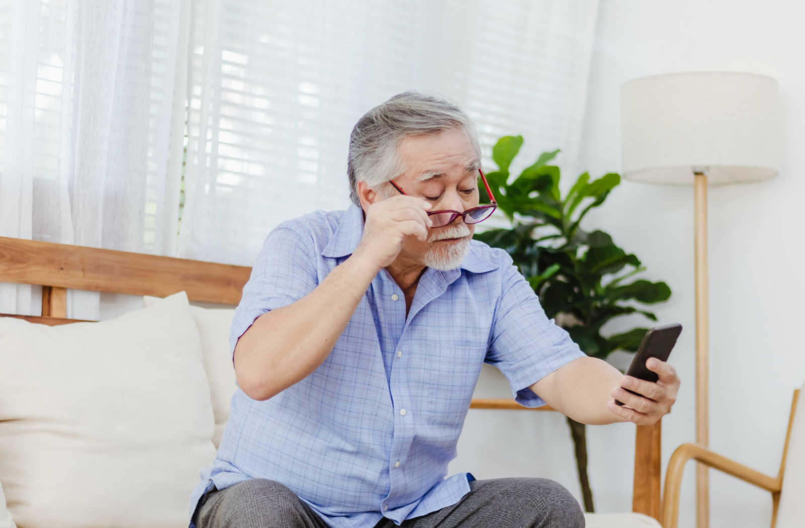 An elderly man in a blue shirt, sitting on a sofa, lifting his glasses with one hand while holding the phone with the other in an attempt to read more clearly.