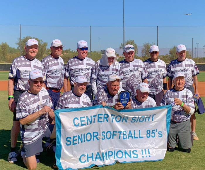Group of senior men in matching jerseys pose on a softball field, holding a trophy and a banner that reads 'Center for Sight Senior Softball 85's Champions.'